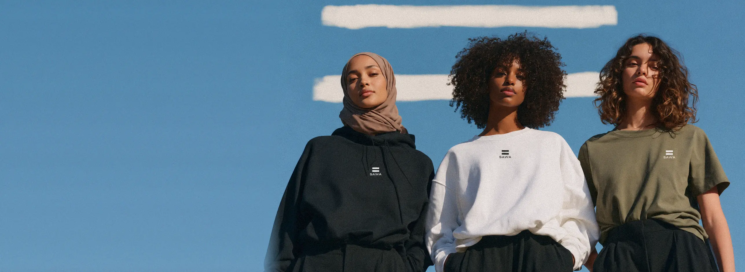 Three women wearing SAWA under a blue sky: black hoodie and taupe scarf, white sweatshirt, olive oversized t-shirt