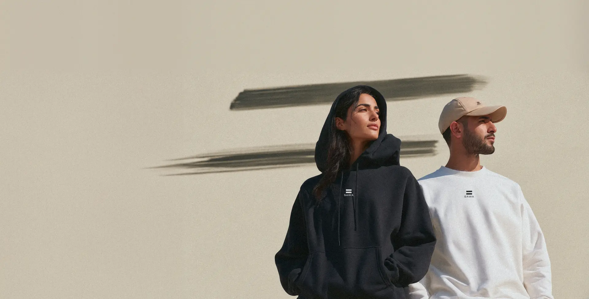 A woman in a black SAWA hoodie with the hood up and a man in a white SAWA sweatshirt and sand cap, in front of a light wall marked with two black brushstrokes